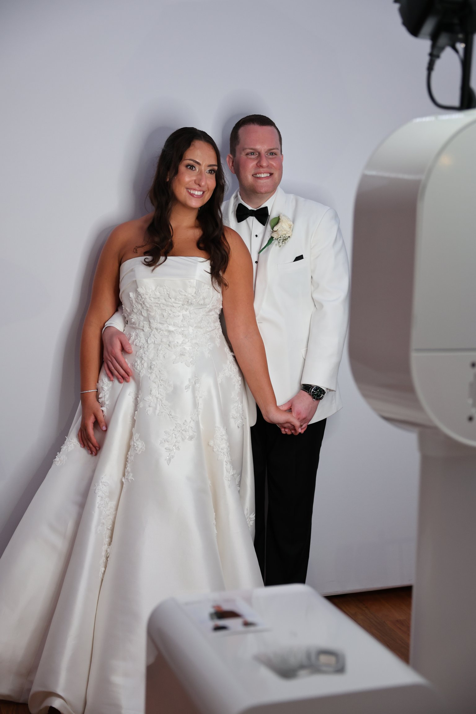A couple in a wedding dress and tuxedo standing beside a white photo booth