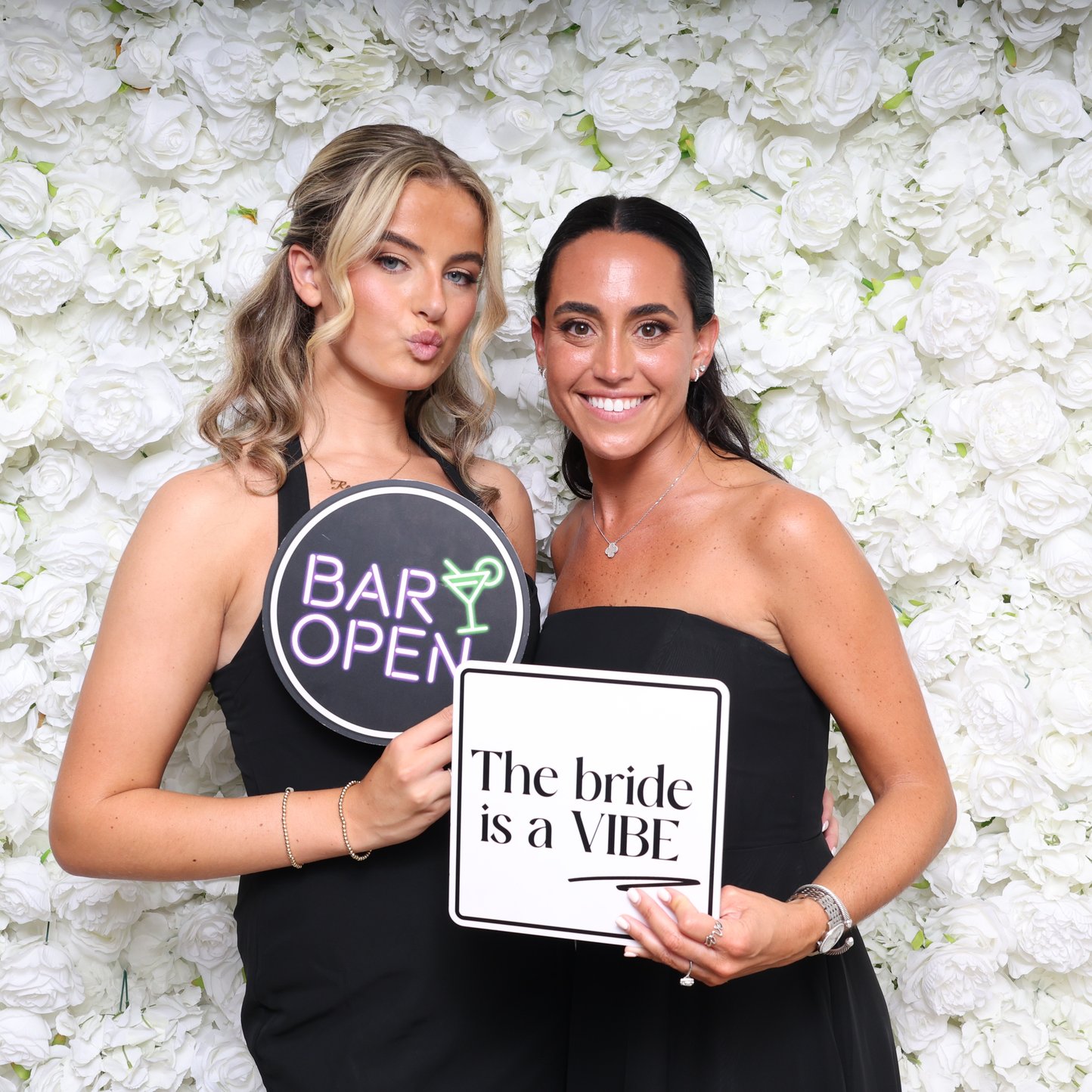 Two women holding hand-held sign props in front of a white flower wall