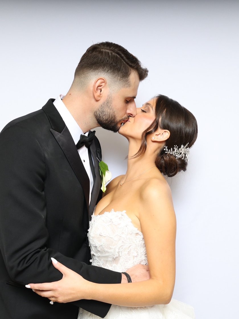 A couple in a tuxedo and wedding dress kissing in front of a white backdrop